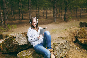 Naklejka na ściany i meble Young smiling beautiful woman in casual clothes with headphones sitting on stone using tablet pc computer in city park or forest on green blurred background. Student lifestyle, leisure concept.