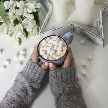 Cocoa with marshmallows in female hands on white background flowers, candles, top view