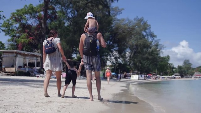 Mom and Dad with backpacks behind, holding his son by the arms and walking along the beach at sea, the daughter sits on his father's shoulders. slow motion, 1920x1080, full hd