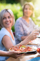 Close-up on a hand serving a dish of dry ham and cheese to friends gathered around a table in a garden to have fun together.