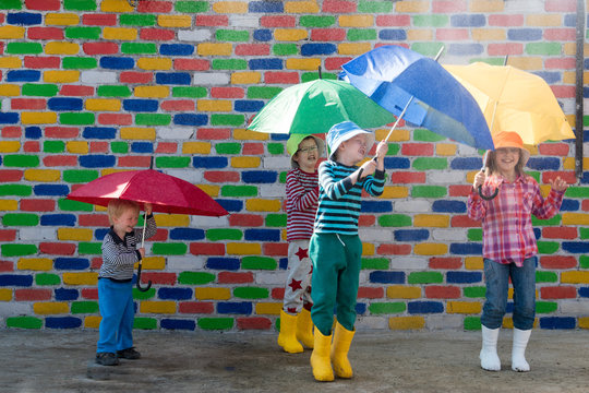 Happy children in colorful hats are sheltered from the rain under multicolored umbrellas in front of mottled brick wall. Fun during the summer holidays. Boys and girl have funny time. Copy space
