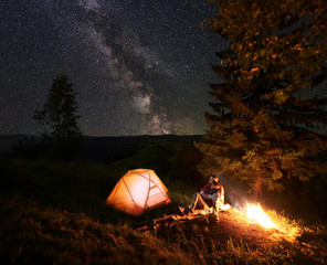 Fototapeta na wymiar Romantic couple hikers sitting on log beside illuminated orange tent, hugging and admire scenery of bright starry sky and Milky way. Pair having a rest by campfire at camping near forest in mountains