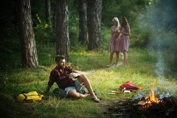 Camping in forest. Man looking at fire while drinking tea. Guy sitting on grass on sunny meadow. Two barefoot girls walking on path.