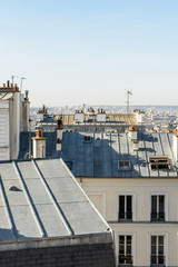 Open view over the rooftops of Montmartre district in Paris with typical zinc roofing, skylights, chimneys and TV antennas under a clear blue sky.
