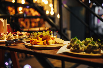 Snacks on a wooden shelf. Different cheese on a plate, grapes. In the background light bulbs. Yellow light. Snacks at the party
