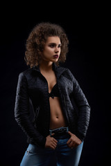 Sexy woman posing in black jacket and blue jeans. Looking beatiful,confident, charismatic. Having pretty face features, many little curles and light day make up. Studio black background.