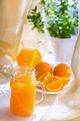 Juicy orange juice in a glass and jug and ripe fruit orange on a white table near the window, the concept of breakfast on a sunny summer day