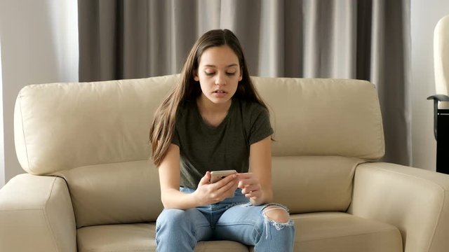 Young teenager browsing through her smartphone on the couch in the living room while her oldest sister is coming in the back and covers her eyes.