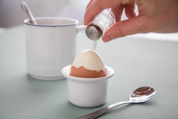 Weibliche Hand würzt gekochtes Ei im Becher mit Salzstreuer beim Frühstück