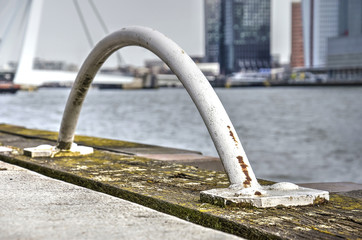 The white steel arch indicating an emergency ladder mounted on a wooden beam on the quay of the river Nieuwe Maas in Rotterdam with parts of Erasmusbridge and Kop van Zuid blurred in the background