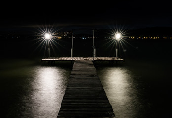 swimming pier on a lake at night with two lights burning bright over shimmering water and the lights on the opposite shore glowing in the distance