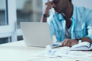 Tired Indian man sits behind laptop in office. Ideological crisis. Crumpled sheets.