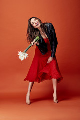 Beautiful smiling brunette woman dressed in red lace dress, black leather jacket and beige heels posing with bunch of daffodils in studio with orange background