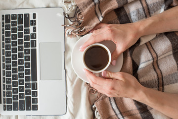 A cup of coffee in the hands of a girl and a laptop on a brown plaid. Top view, morning concept