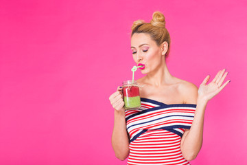 Portrait of funny young woman wearing bright outfit, holding and drinking tasty green smoothie milkshake on pink background with copyspace
