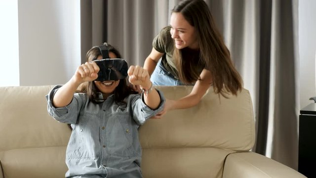 Woman enjoying virtual reality with a VR headset sitting on the sofa while her sister is beside her