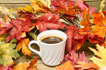 A single hot cup of coffee encircled by leaves in autumn colors on a wooden surface.