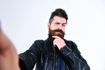 Man with beard and mustache on strict face looking at camera. Macho wears leather jacket, white background. Masculinity concept. Hipster looks serious and thoughtful while taking selfie photo.