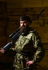 Fototapeta premium Gamekeeper concept. Man with beard wears camouflage clothing, carries rifle on shoulder, wooden interior background. Hunter, brutal hipster on strict face with gun ready for hunting.