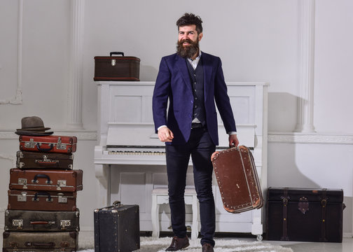Man, traveller with beard and mustache with luggage, luxury white interior background. Baggage delivery concept. Macho elegant on smiling face stands near pile of vintage suitcase, holds suitcase.