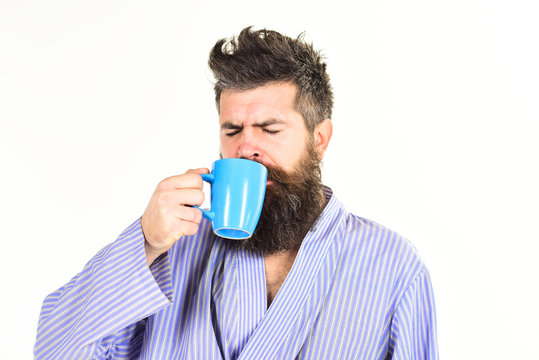Macho in bathrobe drinks tea or coffee with sleeping face. Guy looks drowsy, needs coffee. Morning coffee concept. Man with beard and mustache holds mug of coffee, isolated on white.