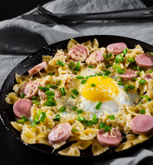 fried eggs with pasta farfalle, sausage and cheese in a frying pan on wooden table