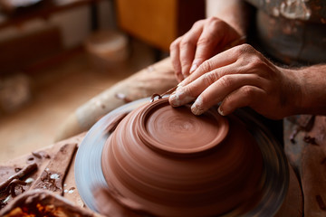 Adult male potter master modeling the clay plate on potter's wheel. Top view, closeup, hands only.