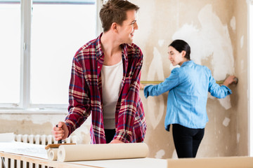 Young man applying adhesive to the back of the wallpaper sheet while his girlfriend is measuring the prepared wall in their new home