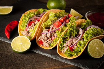 Photo of Mexican tacos with ground meat, beef, beans, onions and salsa on wooden background. Ketchup sauce and lime. A glass o beer in the background.