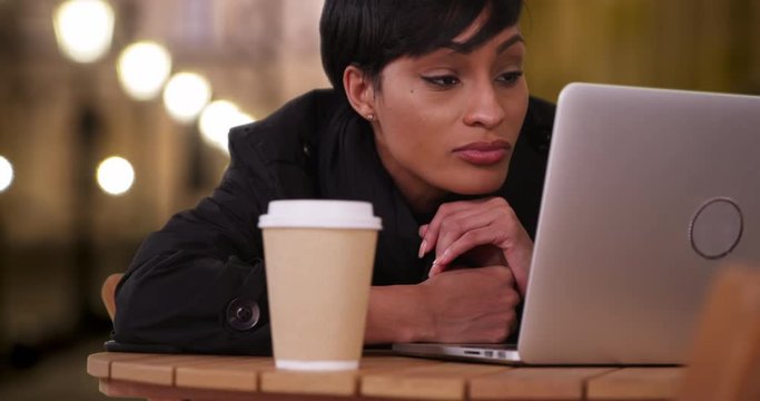 Casual young African female reads something online on her laptop sitting outside at night, Pretty African-American woman at a cafe at night using her laptop computer, 4k