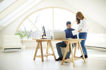 Group of business people working together. Confident businesswoman using mobile phone while sitting at office desk and consulting with young financial assistant businessman. Teamwork at the office. 