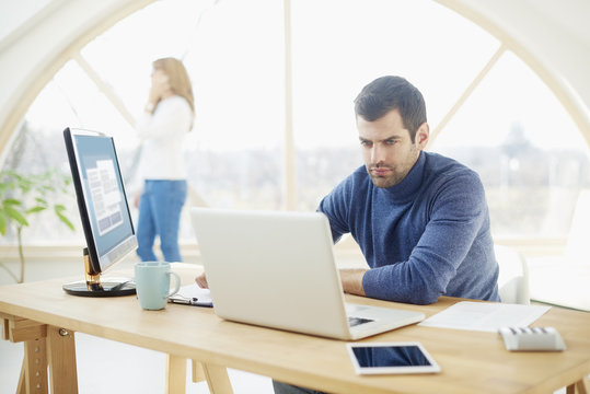 Young man working at the office. Handsome businessman wearing casual clothes while sitting at laptop and typing. Uncrecognizable businesswoman standing at the background. - Powered by Adobe