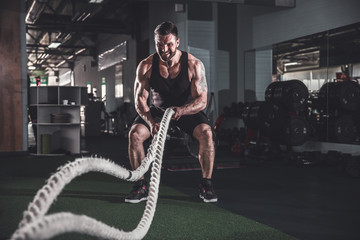 Muscular powerful aggressive man working out with rope in functional training fitness gym