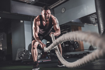 Muscular powerful aggressive man working out with rope in functional training fitness gym