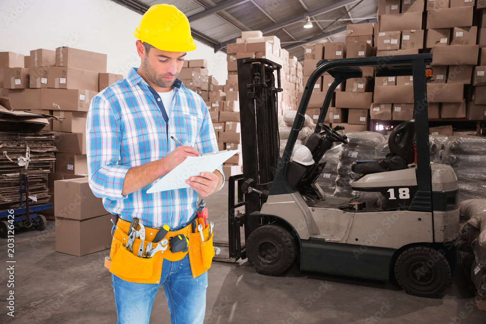 Poster manual worker writing on clipboard against warehouse worker loading up pallet