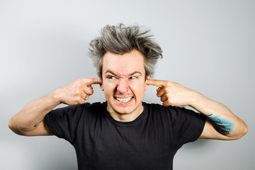 Unshorn and unshaven young guy with piercings on his face, closing his ears with his fingers, on...