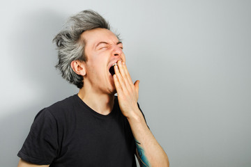 Unshorn and unshaven young guy, yawning, on gray background.