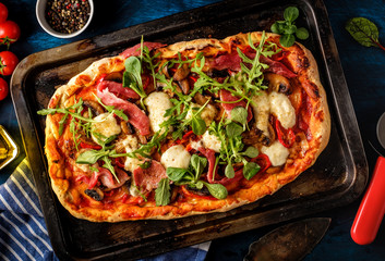 Pizza with prosciutto, mozzarella, mushroom and rocket salad with spices on blue background.