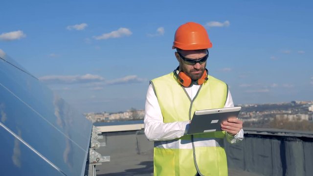 A man types on a gadget on a roof. Worker uses a tablet, while checking how sun panels work.