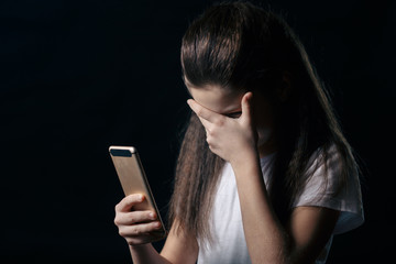 Single sad teen holding a mobile phone lamenting sitting on the bed in her bedroom with a dark light in the background