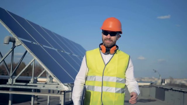 A man with a laptop smiles at camera, standing near solar batteries on the roof. 4K.