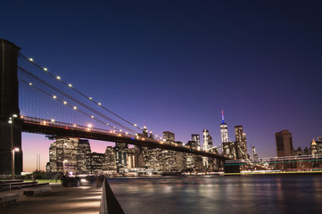 Fototapeta na wymiar Brooklyn Bridge and the illuminated Skyline of Manhattan in the evening with blue sky and smooth water surface shot from Brooklyn side, New York, Usa.