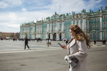 Young traveler woman wearing casual classic coat, holding cellphone, using app, gps, searching for direction while walking in Saint-Petersburg, Palace Square, Winter Palace in spring or autumn time