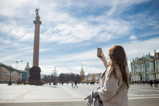 Young traveler woman wearing casual classic coat, holding cellphone and taking photo Alexandrian Column while walking in Saint-Petersburg, Palace Square in spring or autumn time