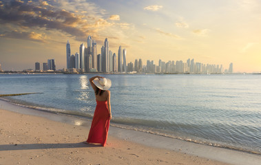 Beautiful woman with a red dress and a white hut is walking on the beach in Dubai. In the background there is the skyline from Dubai Marina