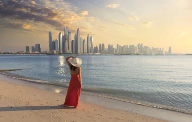 Abwaschbare Fototapete Dubai Schöne Frau mit rotem Kleid und weißer Hütte geht am Strand in Dubai spazieren. Im Hintergrund die Skyline von Dubai Marina