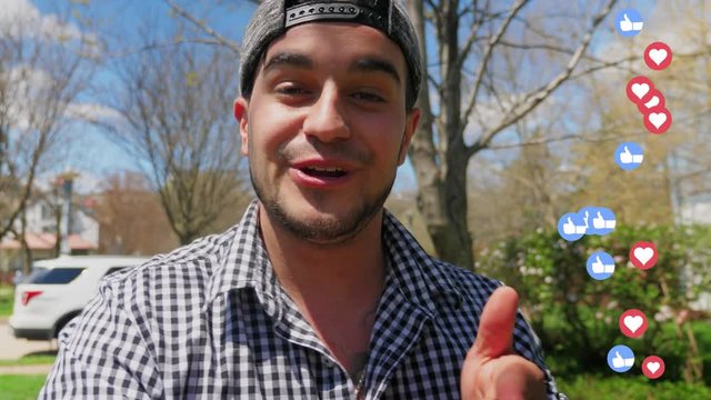 A millennial vlogger wearing a baseball cap backwards talks to his camera while recording a social media video in a residential neighborhood as love and like approval icons fly up on the screen.  	