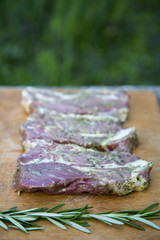 Raw steaks with spices on wooden rustic board against blurred leaves. Ready for cooking. Side view.