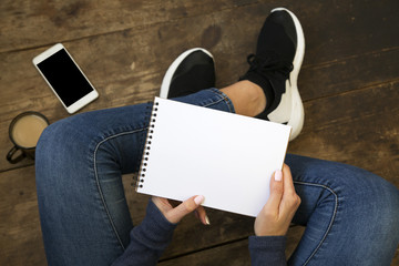Young girl sits on a wooden floor and holds empty notebook in her hands. Smartphone and coffee on the floor. Top view.