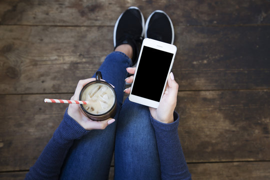 The girl sits on a wooden floor and holds a smartphone with a blank screen and ice latte in her hands. Top view.